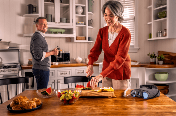 Patient using Optune Lua device while preparing breakfast in the kitchen with a companion.
