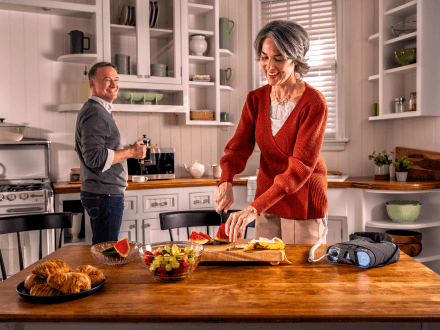 Patient using Optune Lua device while preparing breakfast in the kitchen with a companion.